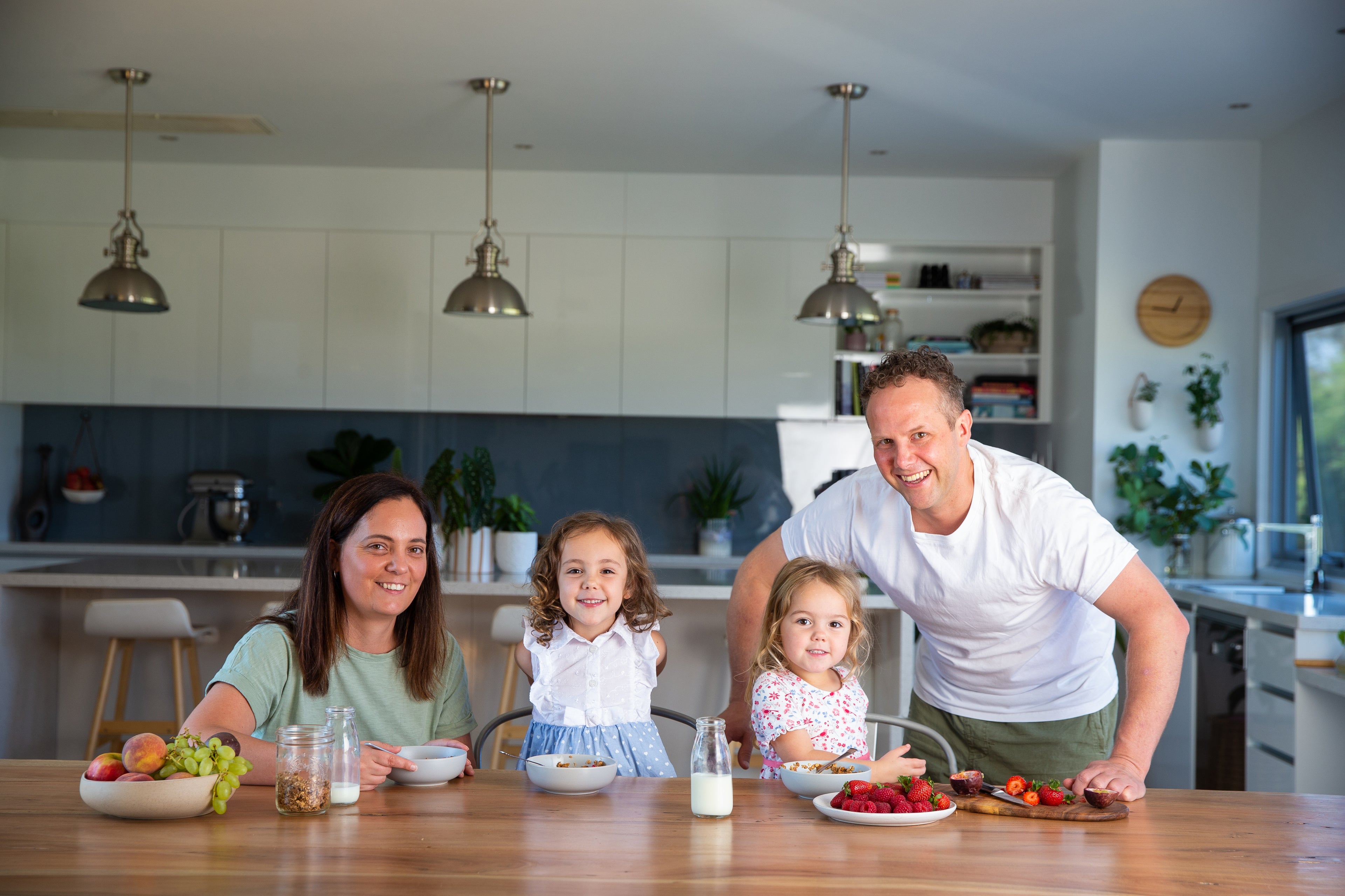 Family of four sitting at a kitchen table with food, smiling at the camera.