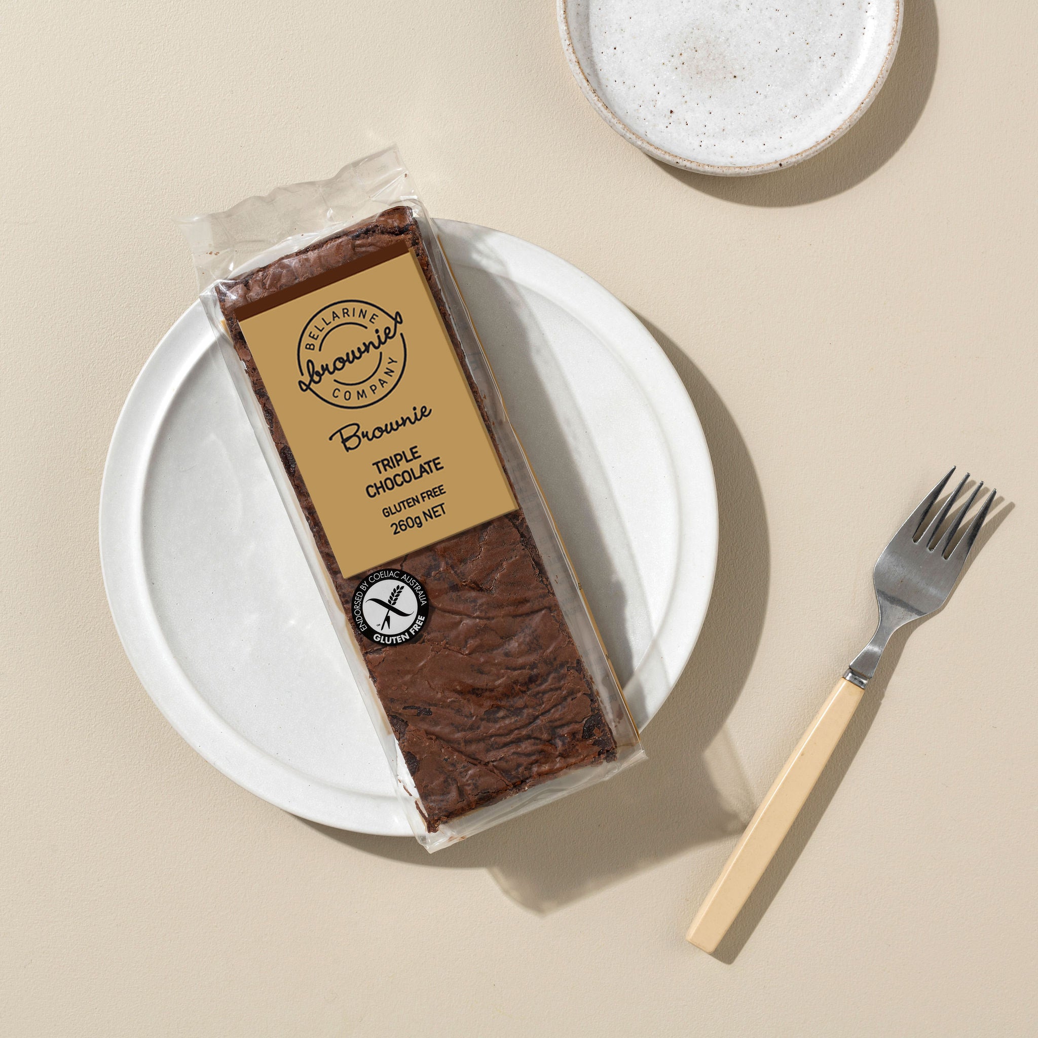 A packaged triple chocolate brownie bar sits on a white plate with a fork beside it on a warm beige surface. 