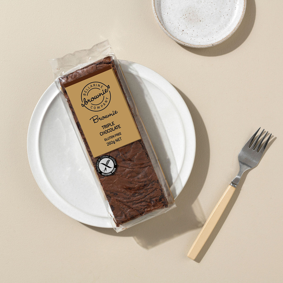 A packaged triple chocolate brownie bar sits on a white plate with a fork beside it on a warm beige surface. 